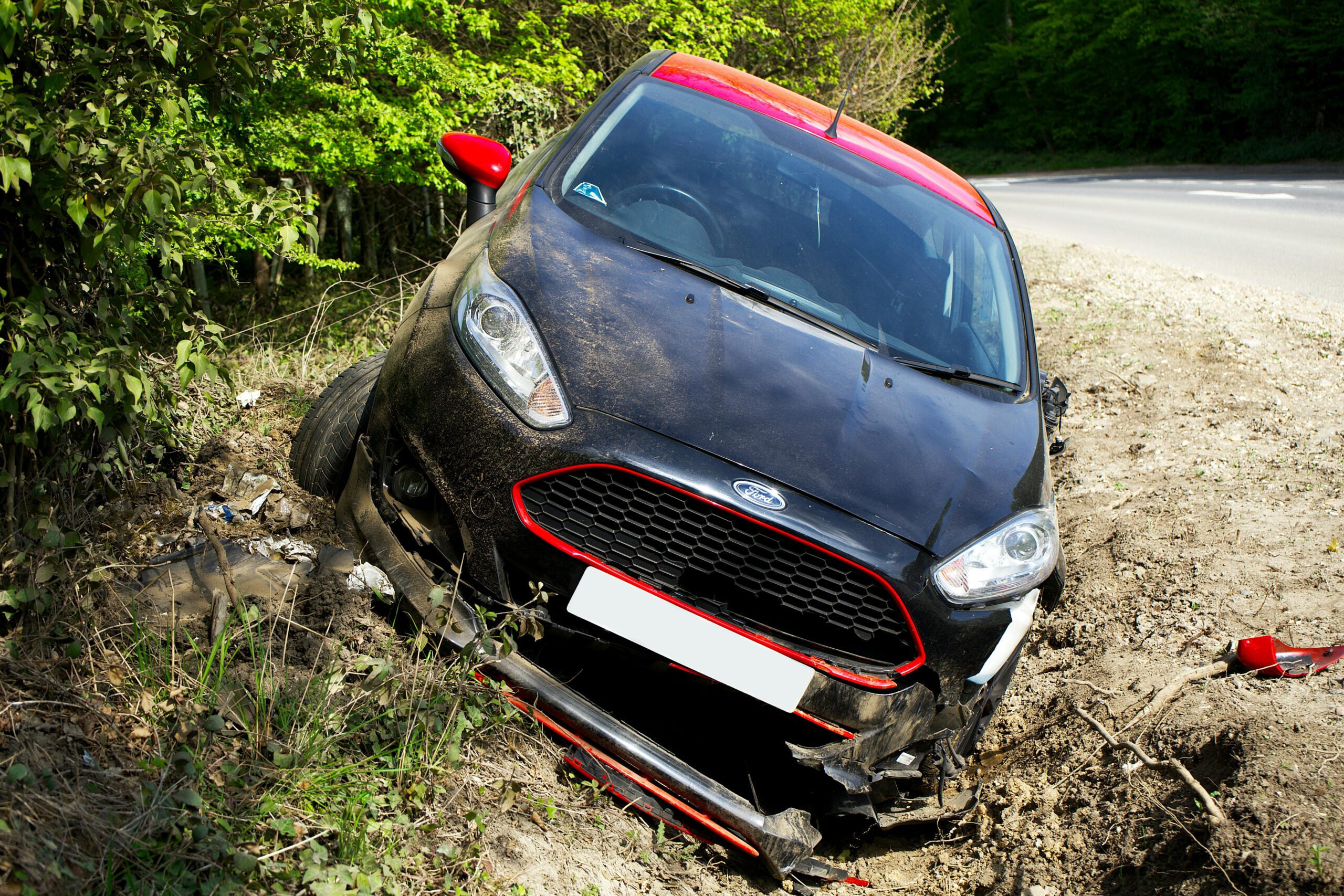 A black car, seemingly a Ford, crashed and damaged in a roadside ditch with surrounding greenery.